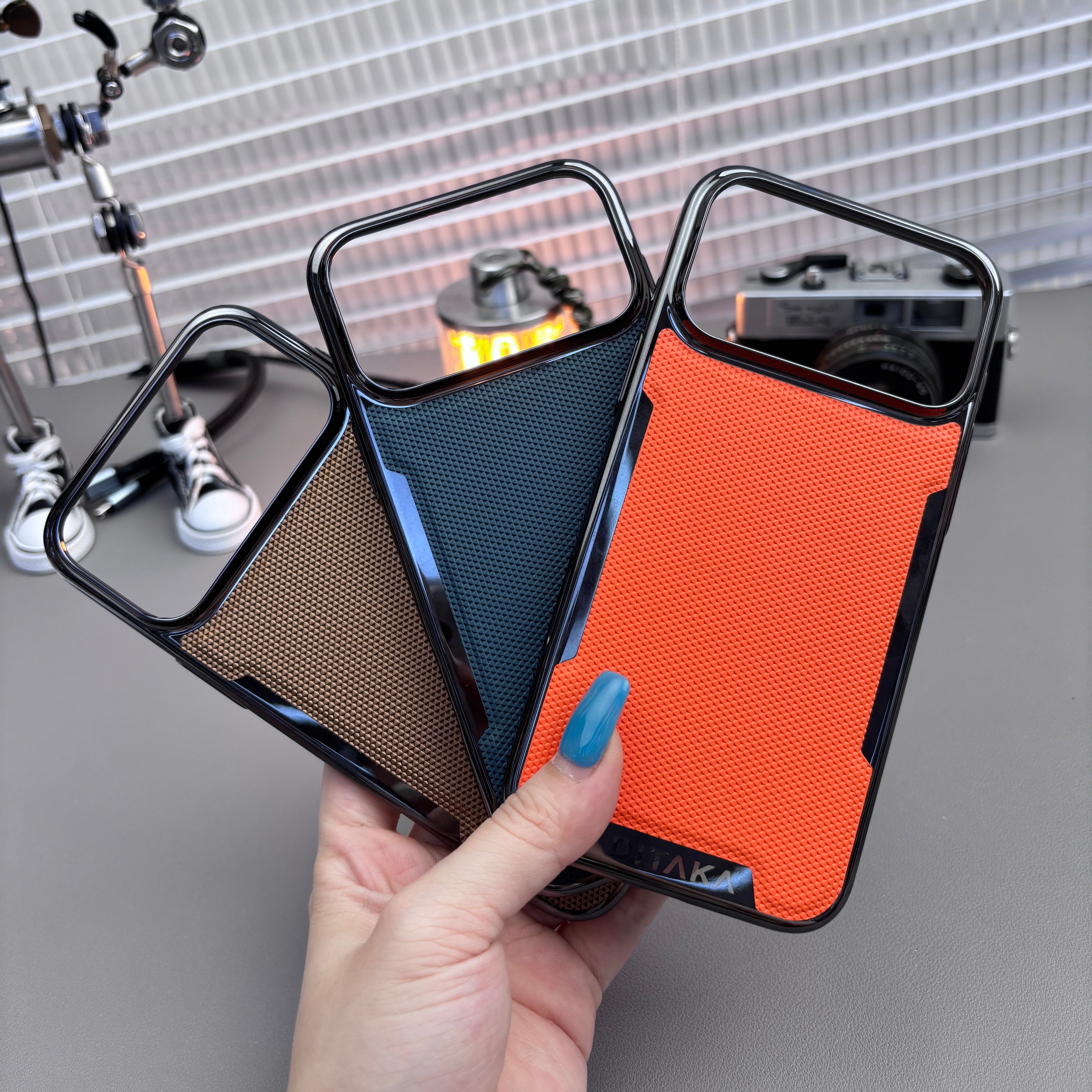 Three phone cases in black, blue, and orange held by a hand against a gray background.